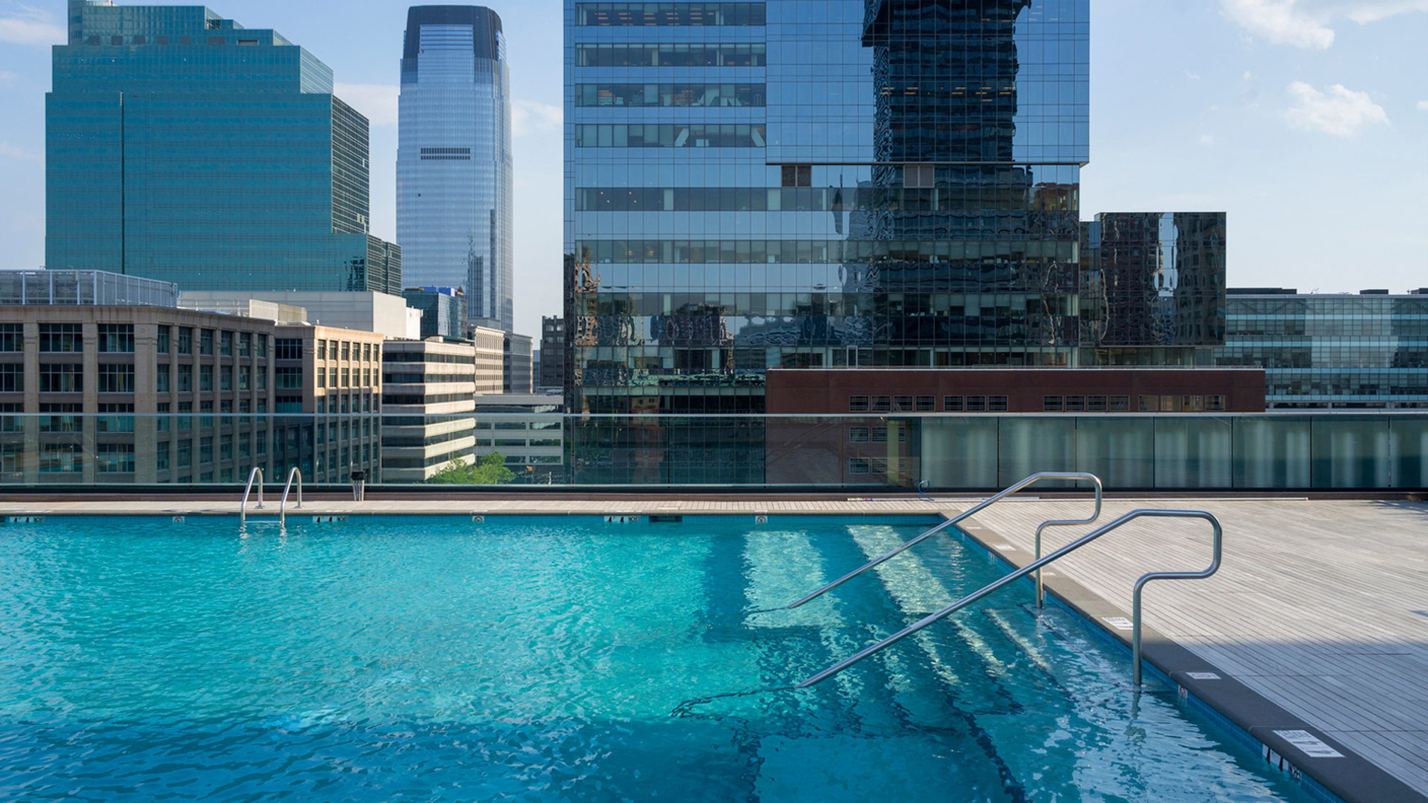 Rooftop swimming pool with a glass safety barrier providing a clear view of the surrounding cityscape. Modern buildings reflect in the water, enhancing the urban oasis vibe. Steel handrails and wooden deck add to the sleek, contemporary design.