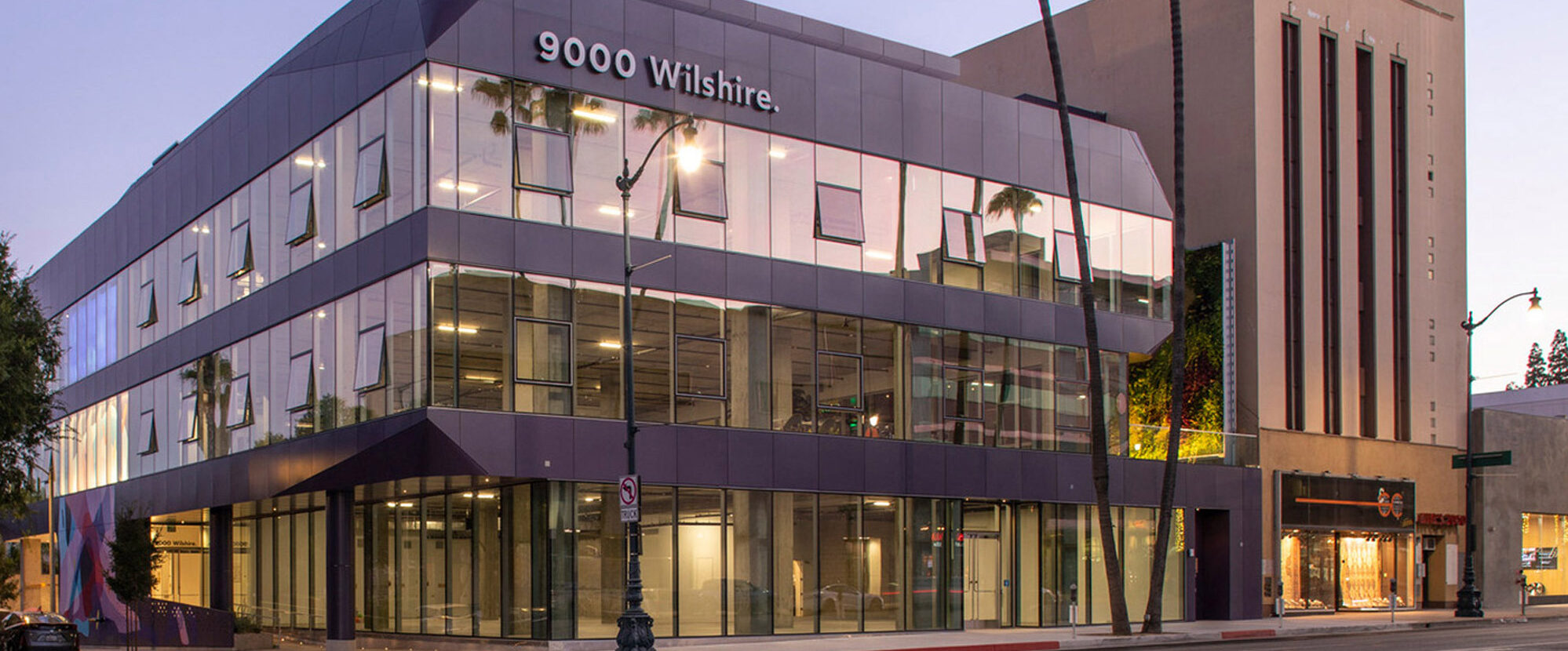 Modern commercial building at twilight with reflective glass facade and cantilevered upper stories. Exterior lighting accentuates the geometric lines and the mix of metal and glass materials used.