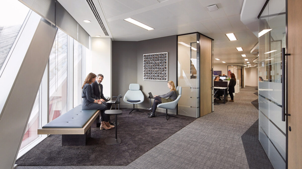 Modern office interior featuring neutral tones with a sleek bench, ergonomic chairs, glass partitions, and art piece, bathed in natural light from floor-to-ceiling windows.