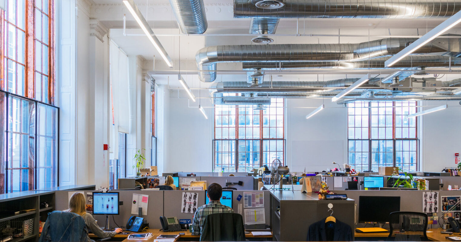 Modern open-plan office space with natural light from large windows with red translucent panes. Features exposed ductwork, white beams, and a mix of seated workspace areas and standing desks, reflecting a contemporary flexible work environment.