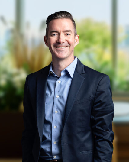 The image captures a man with a friendly smile, sporting a short haircut, wearing a dark suit jacket over a light blue, patterned shirt. He stands with his hands casually at his sides against a backdrop of soft, natural light and blurred greenery, suggesting an office setting with a view of the outdoors.