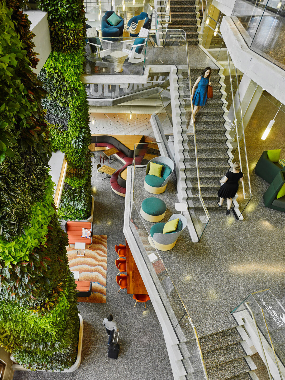 Elevated view of a vibrant multi-level office interior featuring a central spiral staircase with sleek white steps. A vertical garden lines one wall, contributing to the biophilic design. Contemporary furniture in complementary blues and greens pop against the warm wooden flooring and coral upholstered bench seating.