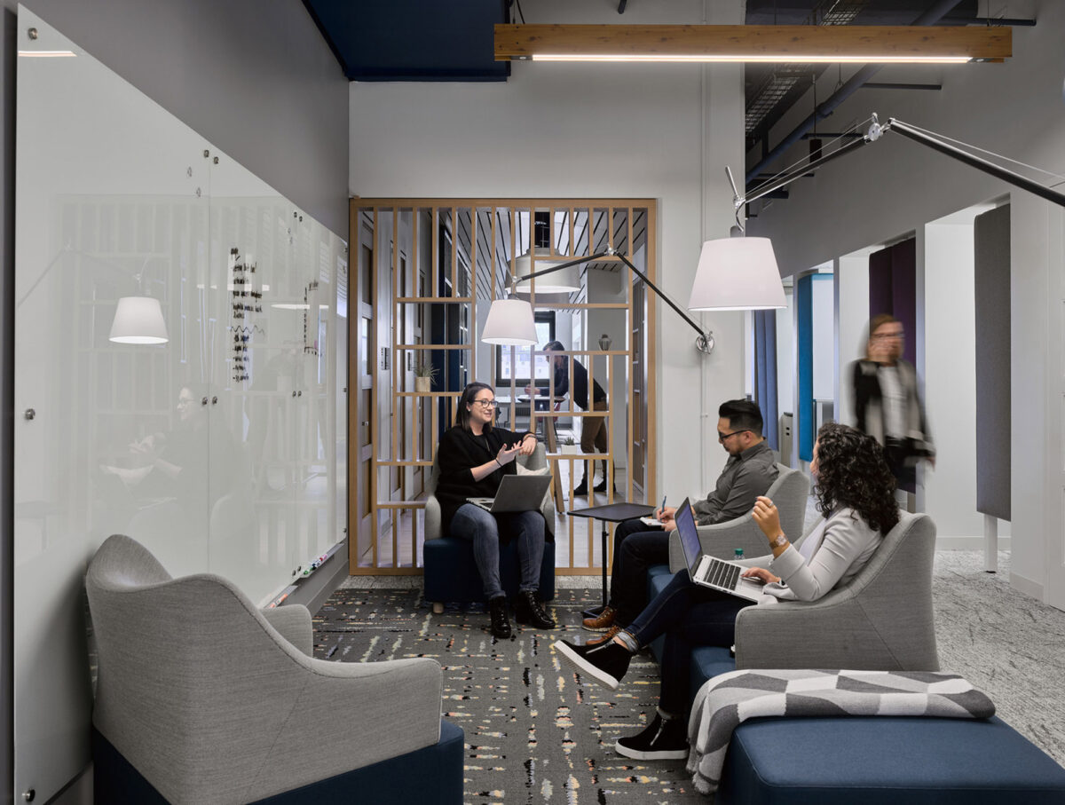 Modern open-plan office space with exposed ceiling beams, sleek gray flooring, and tranquil blue accent walls. Employees engage in collaborative work seated on contemporary armchairs, surrounded by glass partitions and wooden lattice dividers that offer privacy while maintaining visual connectivity.