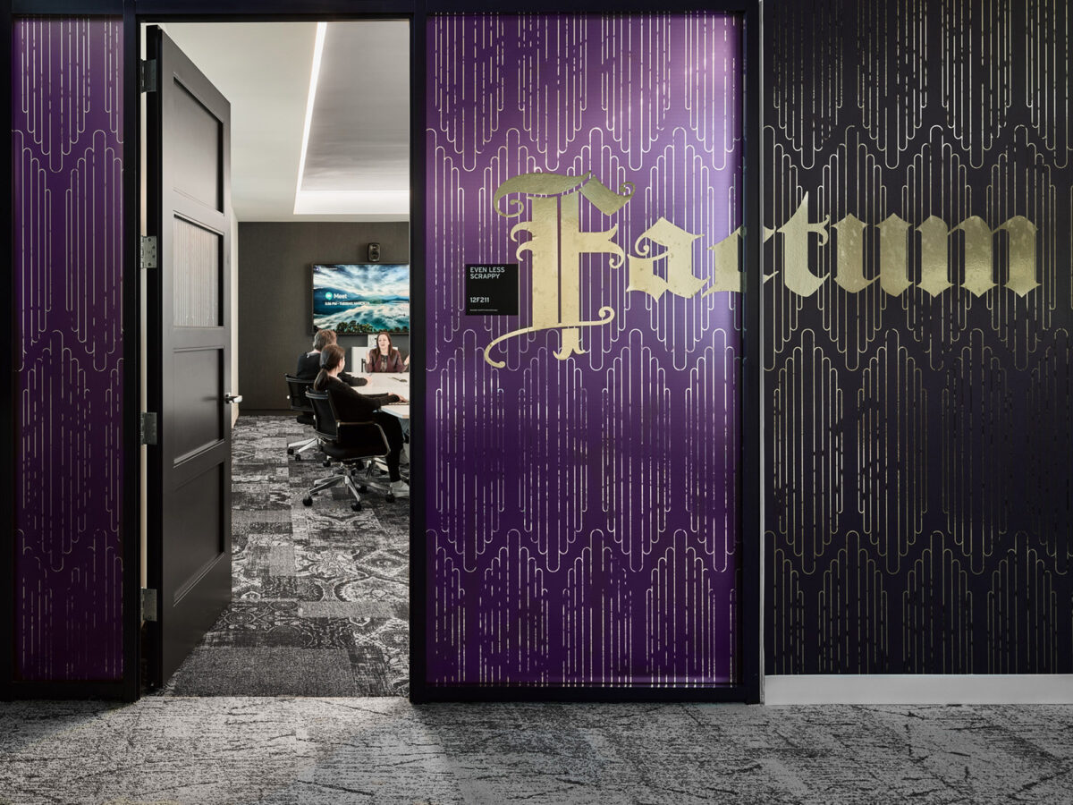 Elegant office entrance with bold purple doors featuring gold lettering, complemented by a circuitry-inspired design. The plush carpet with abstract detailing leads to a workspace with an employee and a coastal artwork.