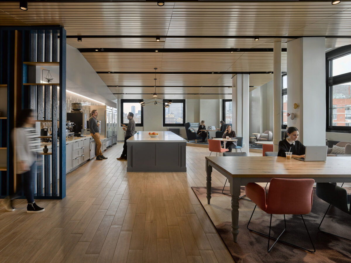 Modern open-plan office space featuring wooden herringbone flooring, ceiling-mounted linear lights, and a central kitchenette with cobalt blue accents. Workers interact and focus on tasks at communal tables and cozy lounge areas alongside expansive windows.