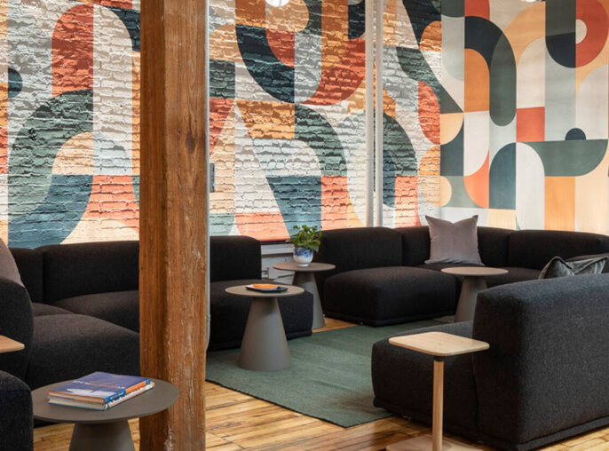 Modern open-plan lounge featuring exposed wooden beams and columns, with a vibrant graphic mural as the backdrop. Earth-tone upholstered modular seating is accented with dark throw pillows, while natural light complements the warm, polished wood floor and planted greenery.