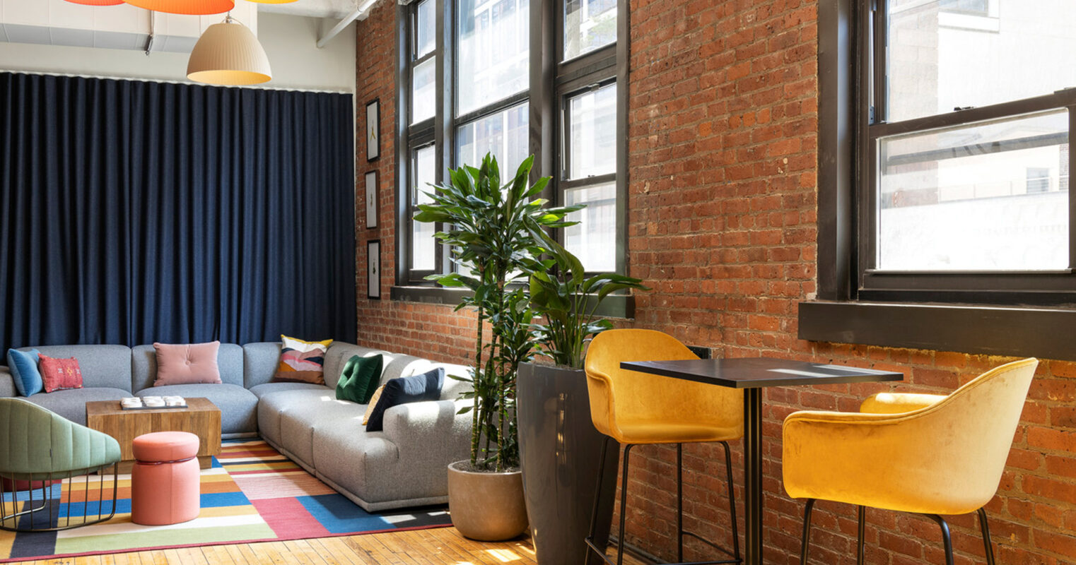 Modern loft living space with exposed brick walls and large windows. Varied colored pendant lighting hangs above a multi-textured seating area. A striped rug anchors the room, flanked by a tall green plant, leading to a cozy nook with bar-height chairs and a wooden table.
