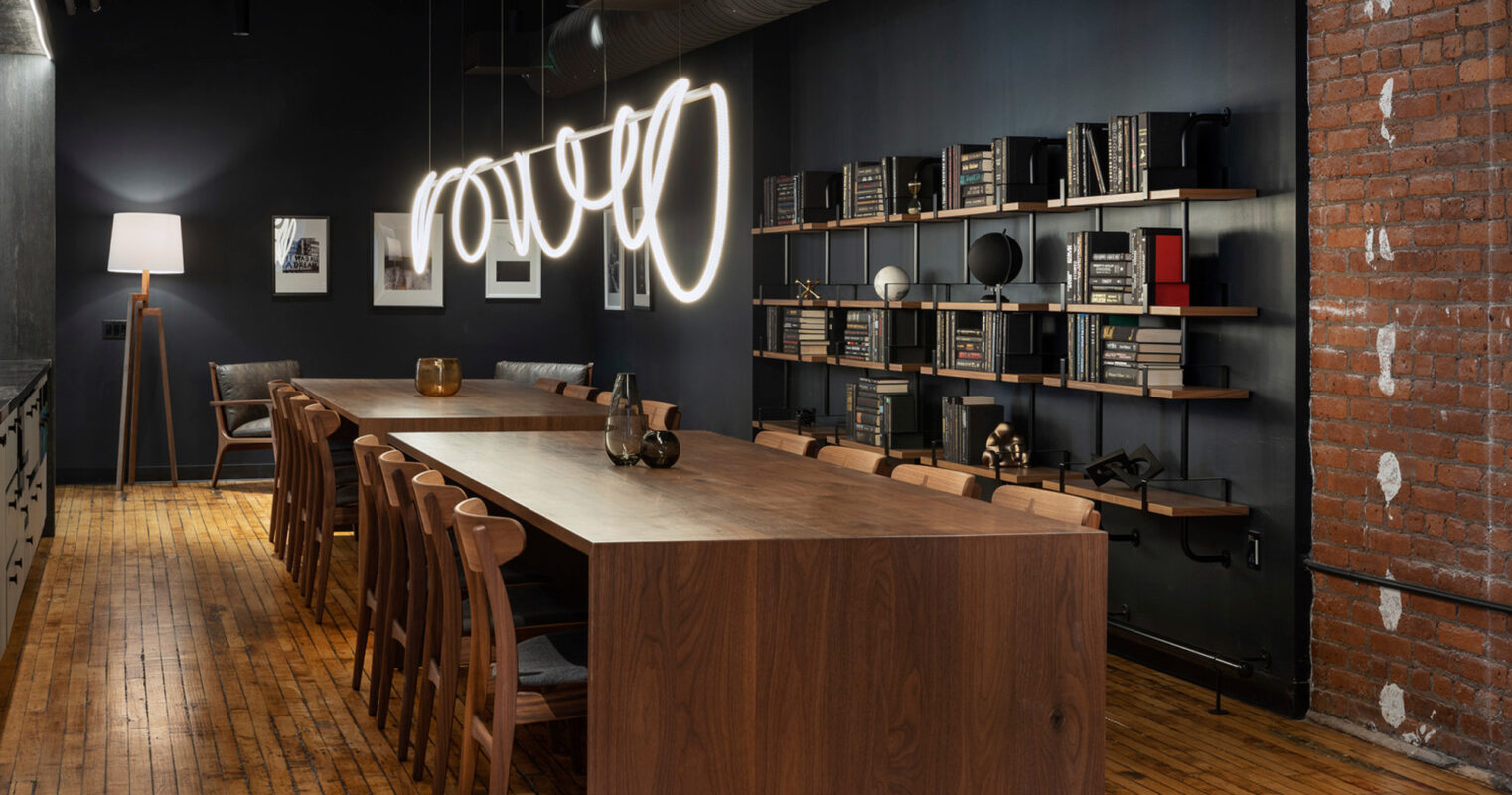 Exposed red brick walls complement the industrial ambiance of this library with a sleek, dark-toned bookcase, warmly lit by a modern neon sign. The elongated wooden table with matching benches creates a communal study space under a cohesive palette of rich, dark hues.