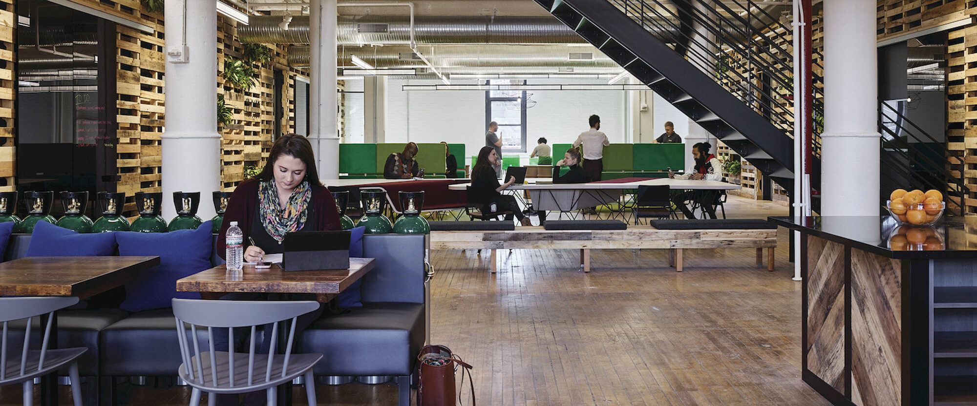 Open-concept office space featuring an eclectic mix of industrial and biophilic design elements, with exposed ceiling pipes, lush greenery partitioning work areas, and hardwood floors complementing contemporary furniture.