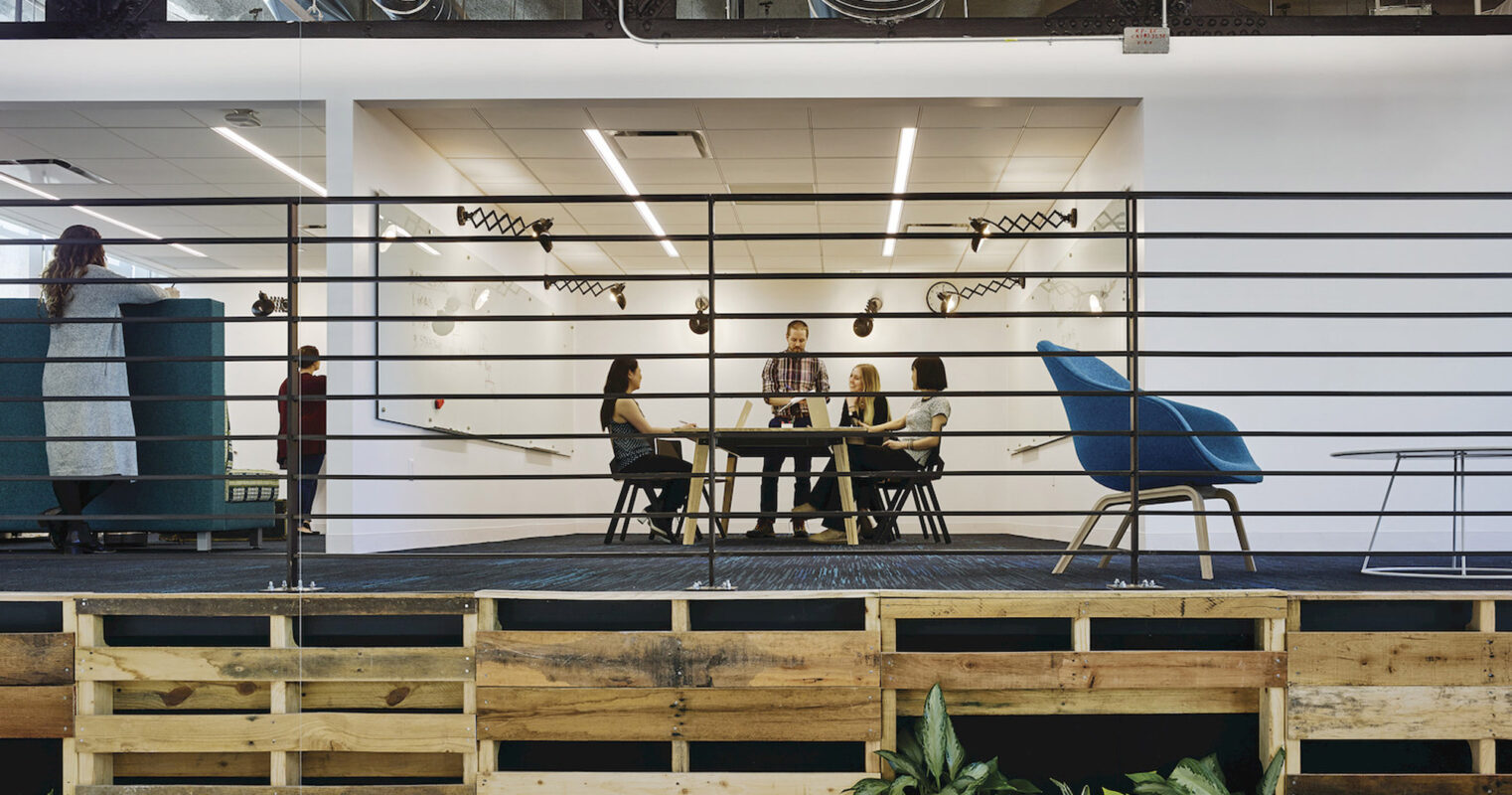 Modern open-plan office space featuring industrial aesthetics with exposed ductwork, natural wood pallet partition, pendant lighting over a sleek conference table, and vibrant blue chairs that add a pop of color to the neutral palette.