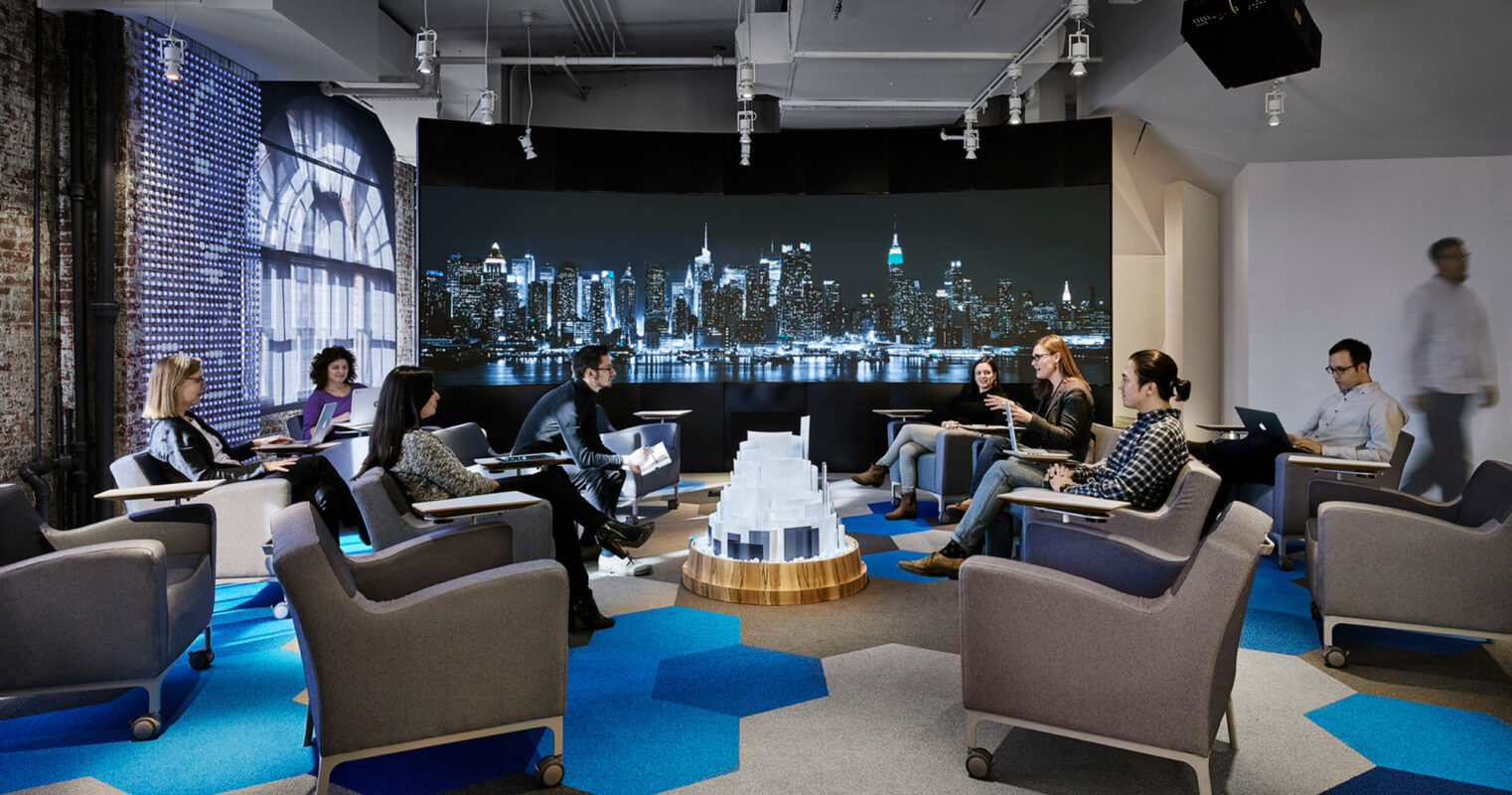 Modern office lounge featuring angular furniture with plush, blue and gray upholstery, complemented by vibrant blue carpet tiles and illuminated by overhead spotlights. A large city skyline mural anchors the space, while a scale model on the central wooden table draws interest.