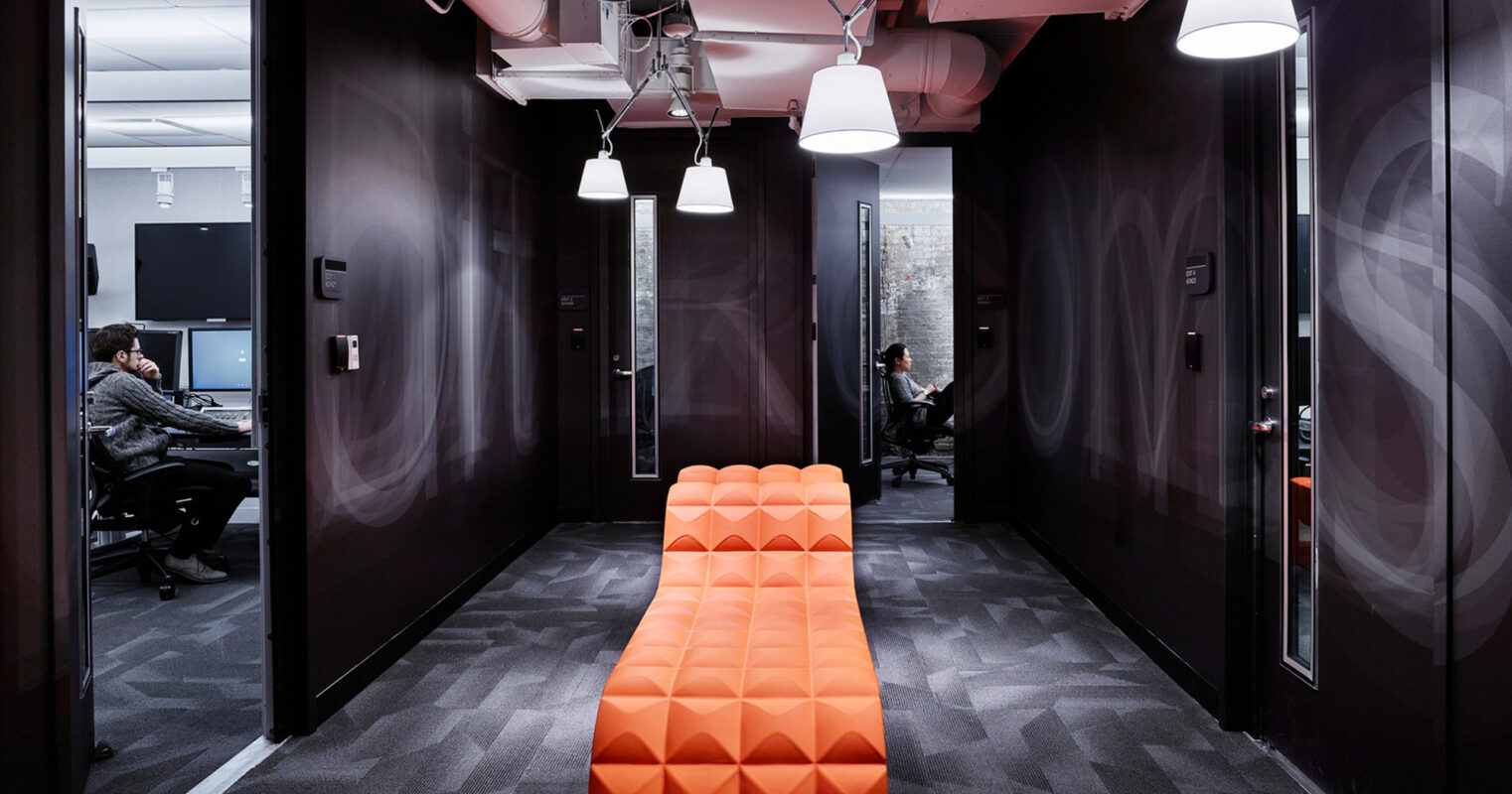 Sleek, modern office hallway featuring geometric patterned carpet, vibrant orange bench with a textured design, contrasting against dark walls and exposed ceiling with angular lighting fixtures.