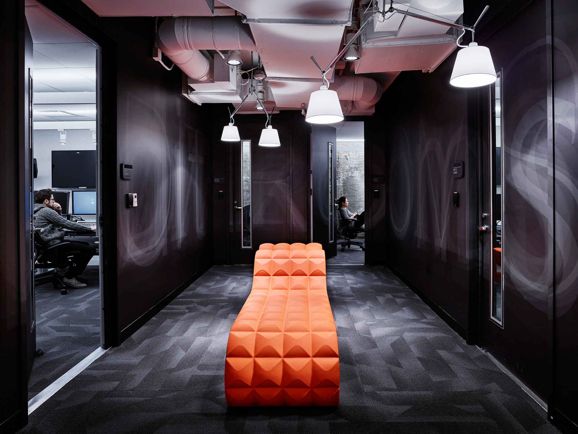 Sleek, modern office hallway featuring geometric patterned carpet, vibrant orange bench with a textured design, contrasting against dark walls and exposed ceiling with angular lighting fixtures.