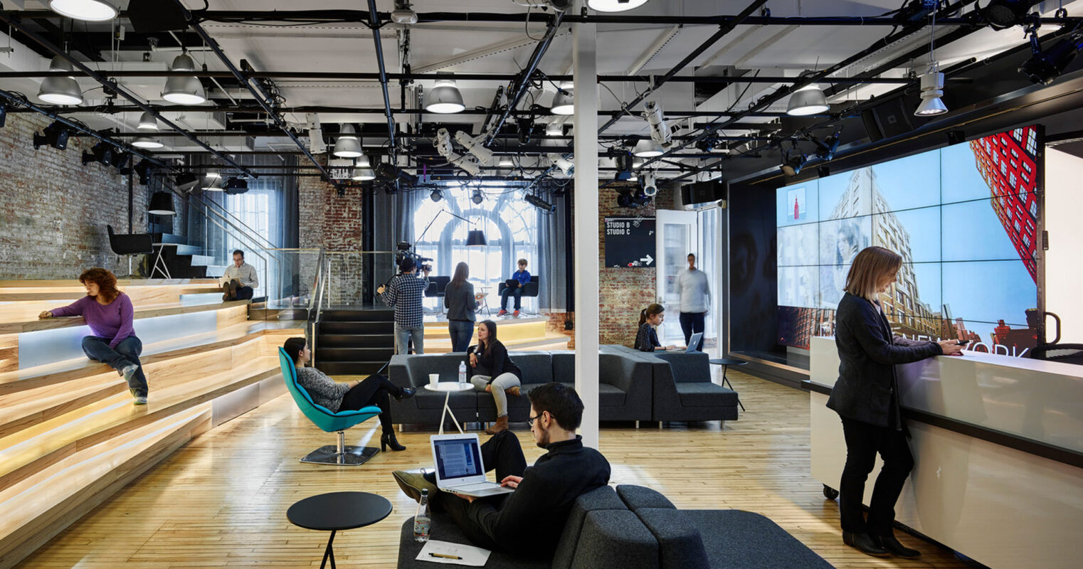 Open-plan office space featuring tiered seating steps, exposed ceiling beams, industrial-style lighting, and a mix of casual workstations with a central interactive digital display. Warm wood tones contrast against a backdrop of brick walls, enhancing the space's modern yet rustic charm.