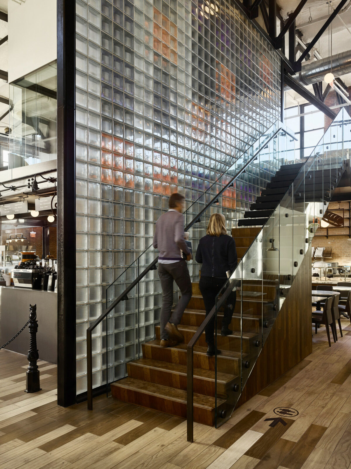 Contemporary restaurant interior featuring a sleek glass block staircase with wooden steps, flanked by a stainless steel and glass balustrade. Industrial design elements like exposed beams add character to the space while patrons ascend the stairs, engaging with the architectural flow.