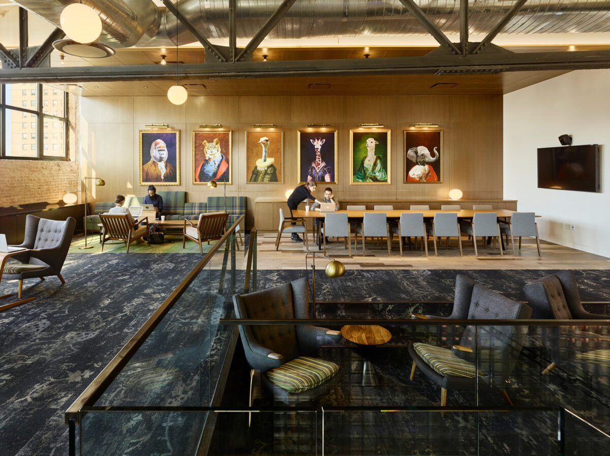 Modern and eclectic workspace featuring an exposed ceiling, rich wooden floors, and a melange of seating arrangements from tufted sofas to communal tables, complemented by vibrant portraiture on the walls.