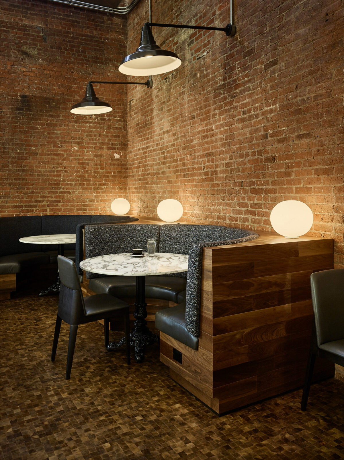 Exposed brick walls complement the warm hardwood flooring in this intimate dining space. Pendant lights cast a soft glow over the marble tabletop, while circular wall sconces add ambient lighting. Two-tone upholstered booths combine comfort with a modern, sleek design aesthetic.