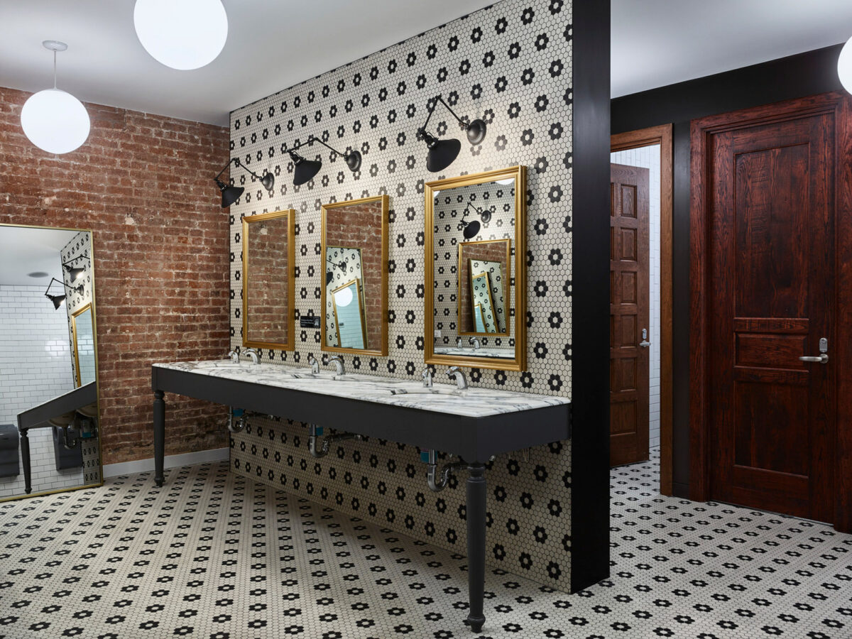 Contemporary bathroom featuring exposed brick walls and a patterned tile floor. Three ornate mirrors hang above a dual sink vanity with industrial style faucets. Contrast is provided by the dark door and light fixtures.