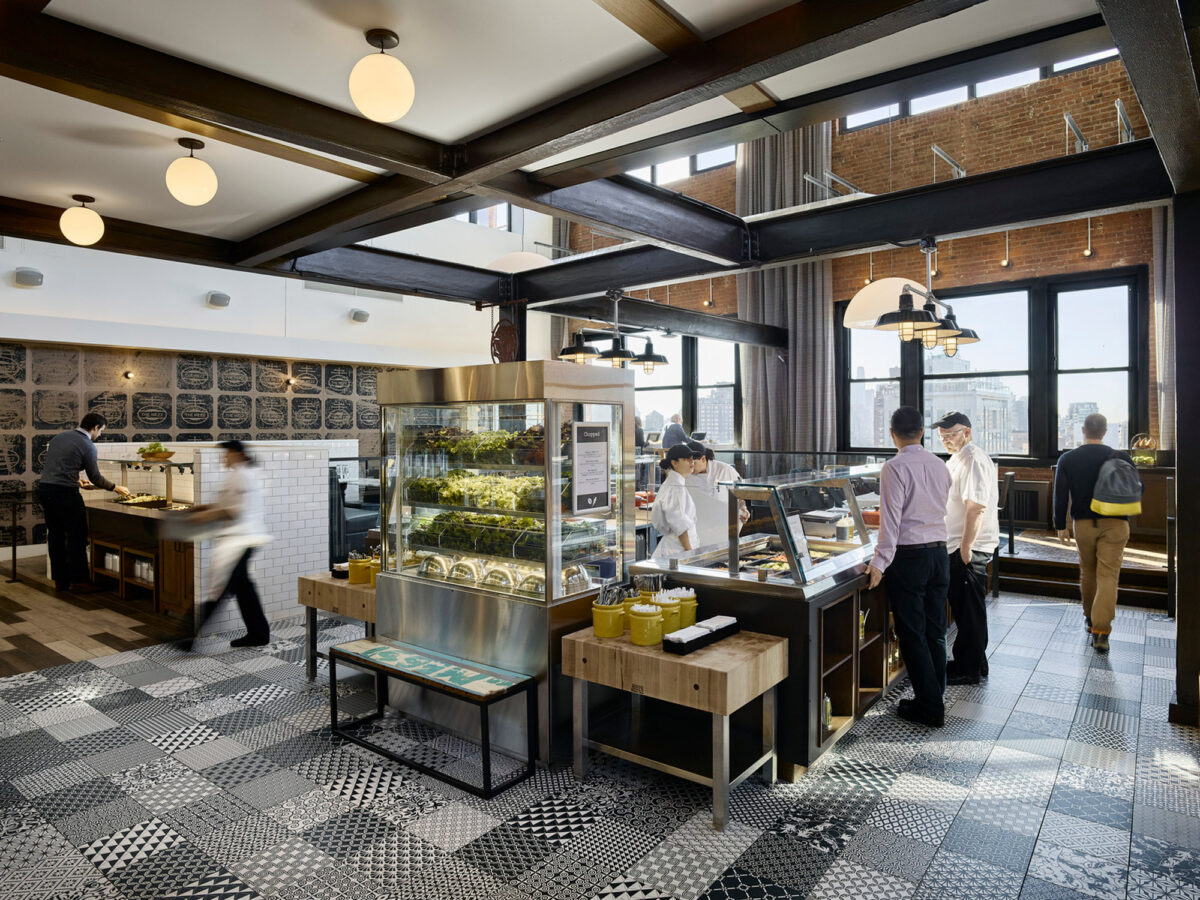 Spacious industrial-style café featuring exposed brick walls, large windows for natural light, geometric floor tiles, and pendant lighting with a central serving area and scattered dining tables.
