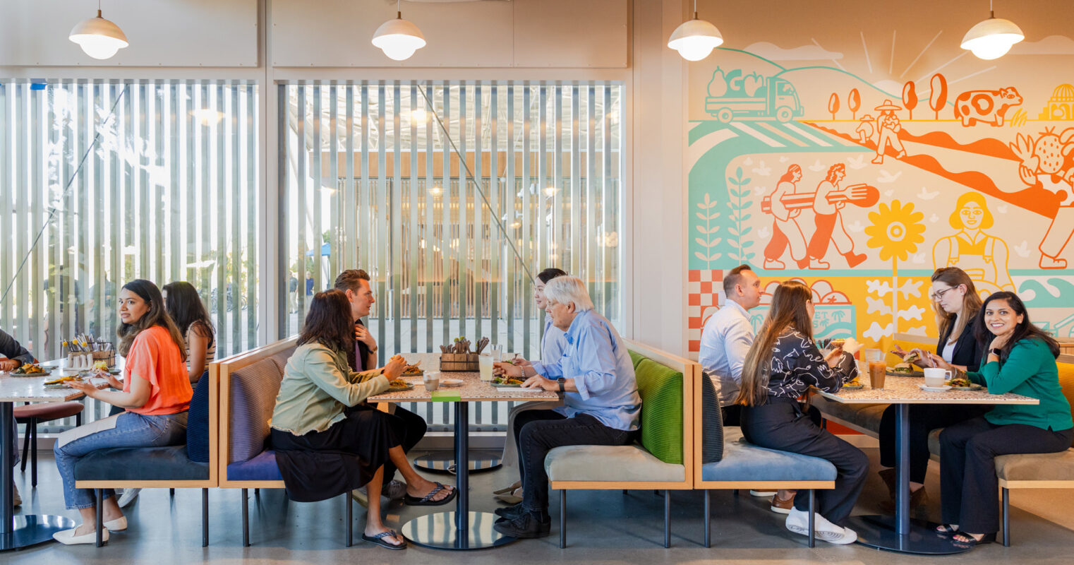 Modern restaurant interior with patrons dining at tables. The space features a vibrant mural with abstract figures, flanked by warm wooden slats and soft, ambient lighting. Comfortable seating contrasts with sleek, minimalistic tables, fostering a casual yet chic atmosphere.
