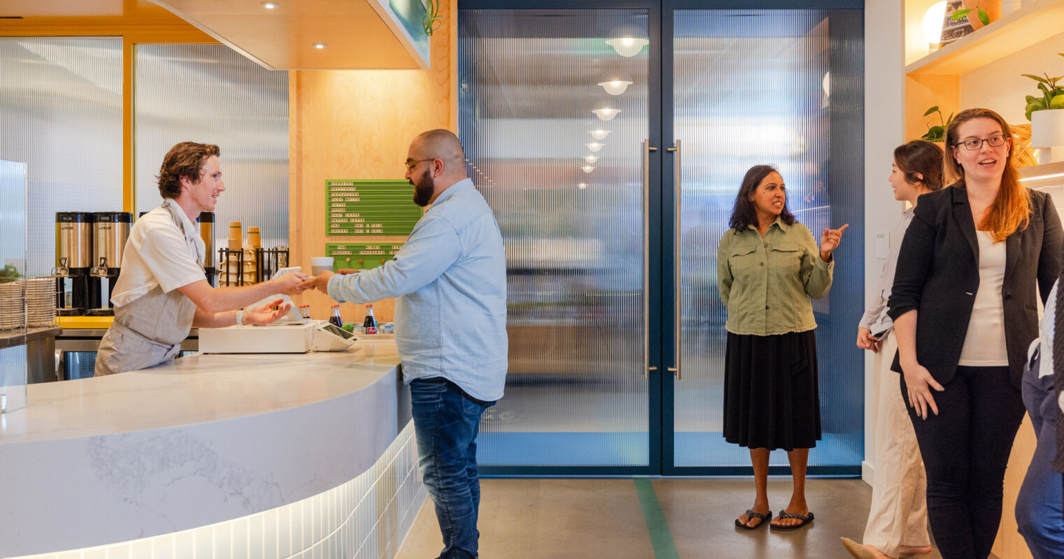 Reception area featuring a curved white counter, blue glass doors, and a warm, wood-accented ceiling. Natural light illuminates the space, enhancing a welcoming ambiance for conversing visitors.