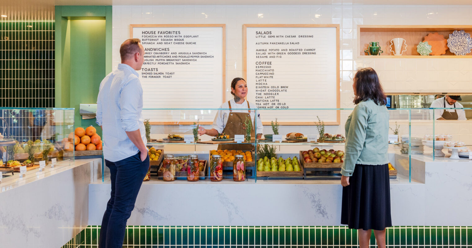 Modern café interior featuring a clean, minimalist design with a pastel color palette, geometric green tile backsplash, and natural wood accents. The space includes a well-lit display case and menu boards in a sans-serif typeface.