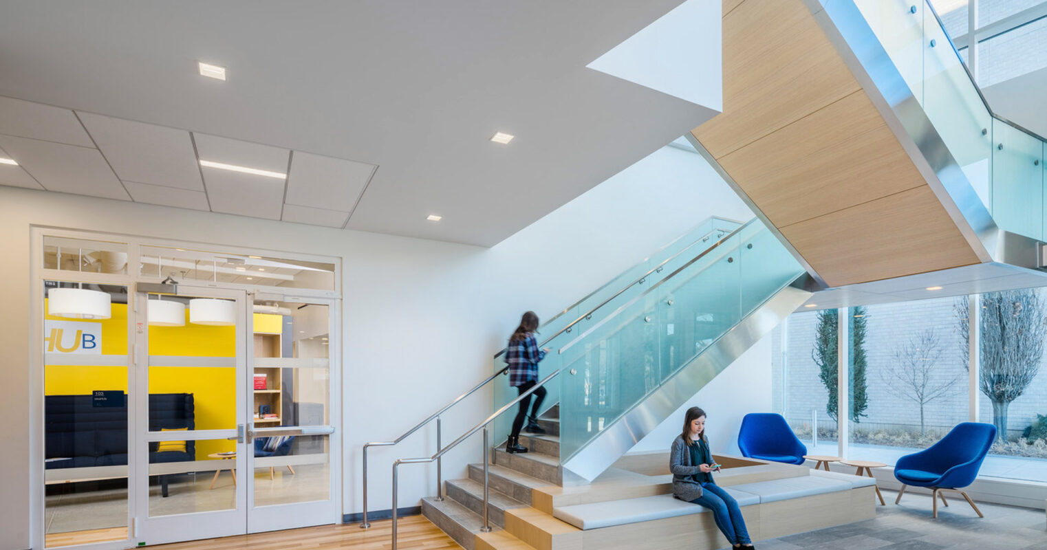 Modern lobby with a soaring glass facade, integrated lighting under the staircase, and a mix of wooden flooring and white walls. Blue mid-century modern chairs add a pop of color beside the expansive glazing overlooking a tranquil exterior courtyard. A person descends the stairs, another is seated using a laptop.