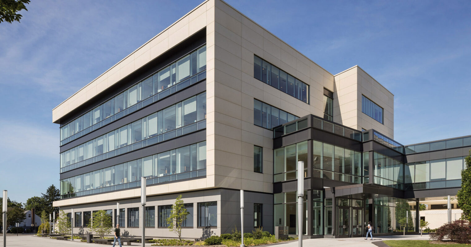 Modern three-story commercial building with an asymmetric facade, featuring a combination of reflective glass windows and pale stone cladding. The ground level’s expansive glazing enhances the indoor-outdoor connection, set against landscaped surroundings.