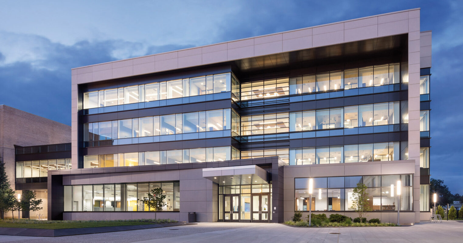 Modern architectural design featuring a multi-level office building with illuminated windows against twilight sky, showcasing reflective glass facades and geometric symmetry with an emphasis on horizontal lines.