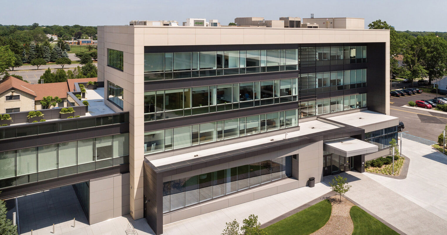 Modern commercial building featuring a sleek facade of reflective glass windows, punctuated by horizontal metal panels, and a protruding entrance canopy with integrated lighting, offering a contemporary architectural aesthetic within a landscaped urban setting.