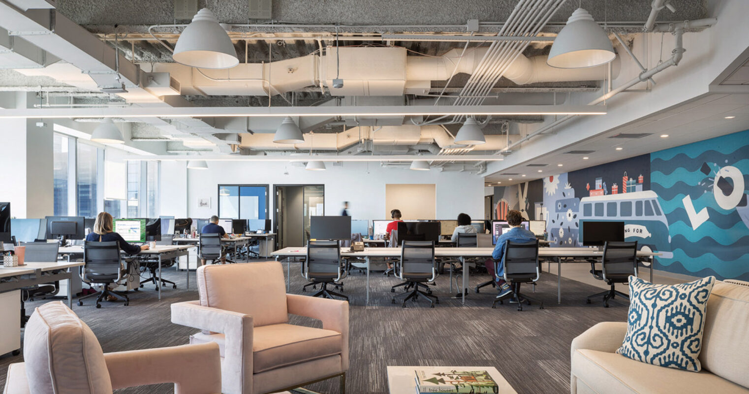 Exposed ceilings with visible ductwork and linear lighting fixtures give this office space a contemporary industrial vibe. Open workstations with ergonomic chairs are arranged for collaborative workflow, while the lounge area features plush seating and decorative pillows, inviting casual meetings. A mural wall injects a playful element into the design, contributing to the overall creative atmosphere. Natural light fills the room, enhancing the blend of functionality and comfort.