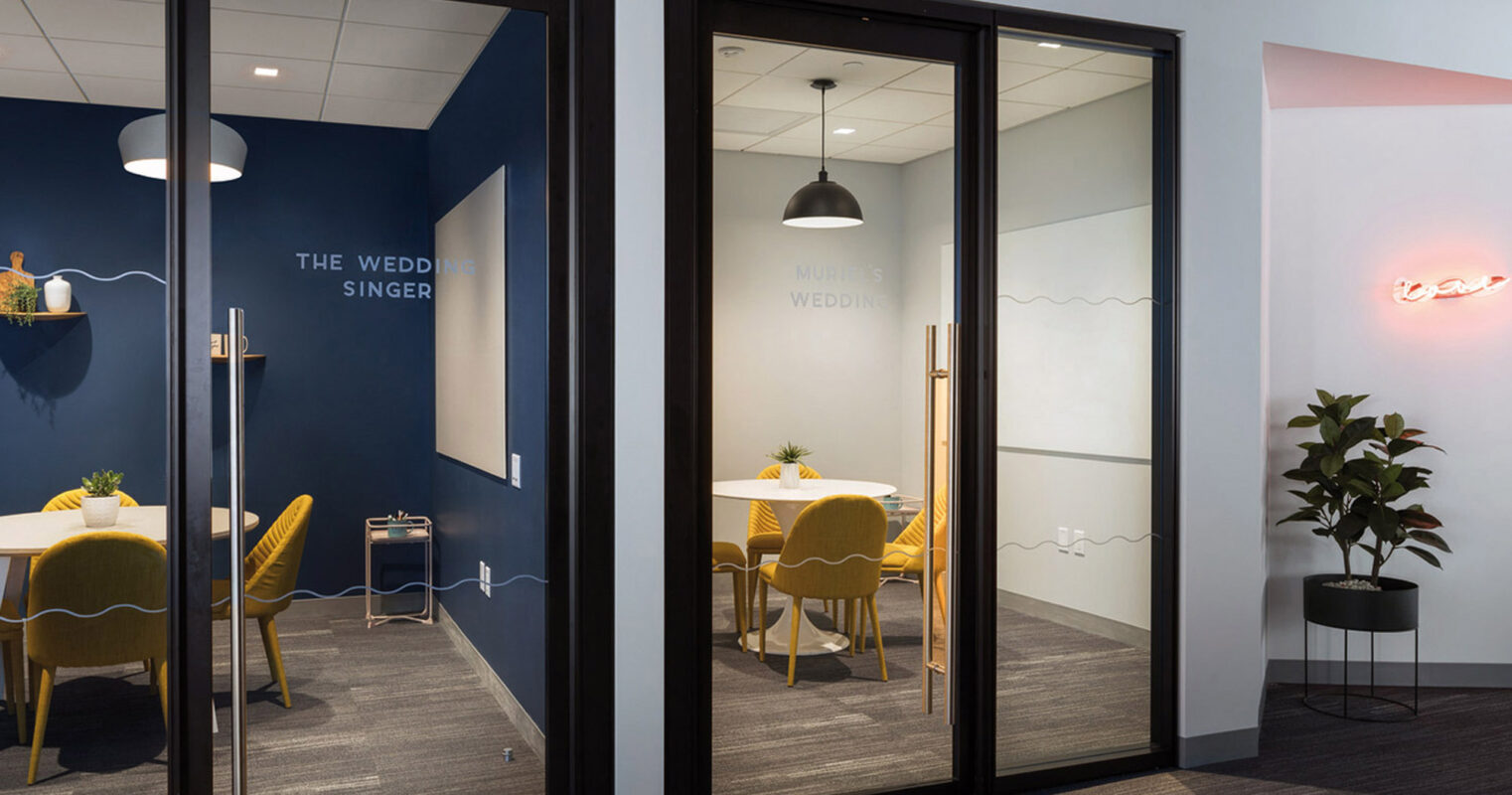 Frosted glass doors open to a modern meeting space with deep blue walls and vibrant yellow chairs, accented by simplistic white lighting fixtures. The room is named "The Wedding Singer," as vinyl lettering on the wall indicates, providing a unique thematic identity. A single potted plant adds a touch of greenery, contrasting with the clean lines and neutral carpeting that anchor the room's aesthetic.