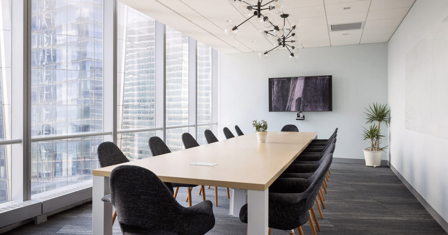 Modern conference room boasting natural light from floor-to-ceiling windows, featuring a minimalist light wood table, charcoal upholstered chairs with wooden legs, and a statement sputnik-style chandelier. A large flat-screen monitor punctuates the pale blue wall, complemented by discreet greenery.