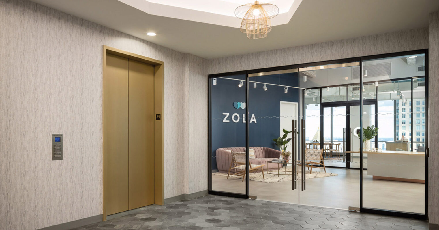Modern commercial lobby featuring a hexagonal-patterned gray tiled floor that transitions to lighter wood flooring inside the glass-enclosed office space. The elevator with satin gold doors complements the neutral textured wallpaper. Overhead, a curved ceiling design with recessed lighting adds depth, while a wireframe pendant light serves as a focal point. Bright natural light fills the workspace, suggesting a high-rise setting with exterior