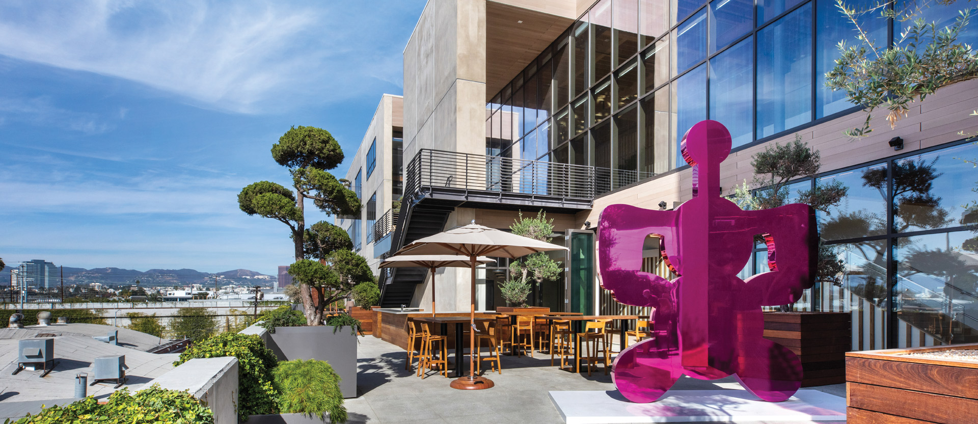 Rooftop terrace with modern outdoor furniture set against a backdrop of architectural glass and clean-lined structures. Purple abstract sculpture adds a pop of color, with greenery accentuating the urban landscape view.