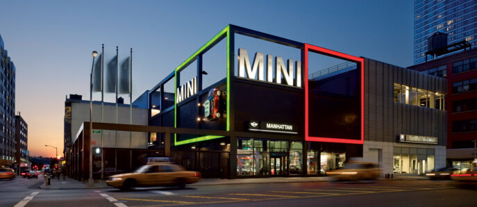 Exterior of a modern car showroom at twilight, featuring vibrant neon-lit framing in green, red, and white, and large glass windows that reveal sleek automobiles and minimalist interior design.