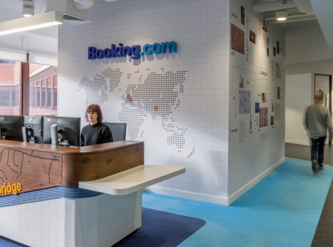 Contemporary office reception area featuring a world map mural on white tiled background. Wood-paneled reception desk with etched city map of Cambridge, against a vibrant blue carpet, enhancing the travel theme. Exposed ceiling pipes add an industrial touch contrasting with the welcoming ambiance.