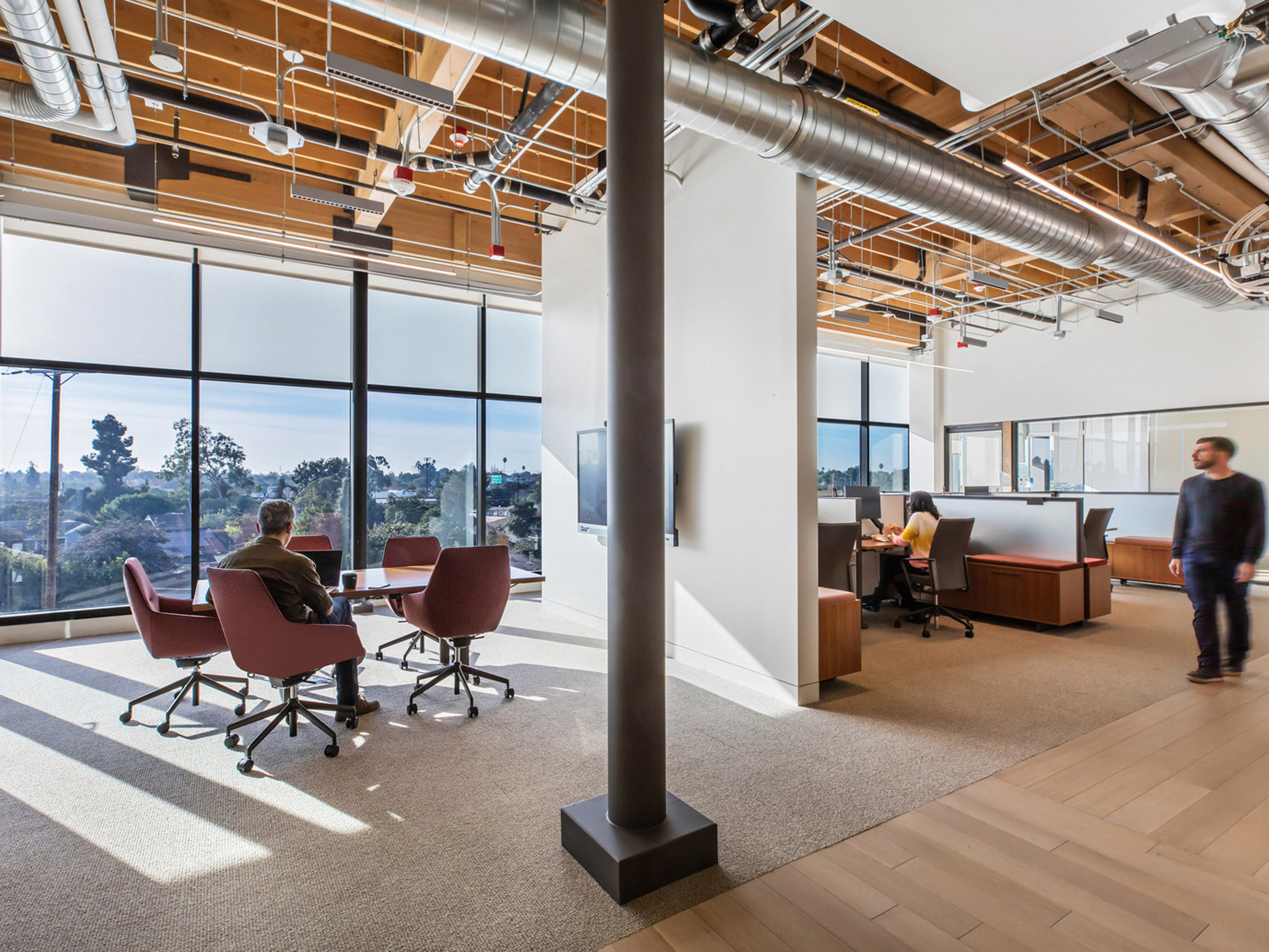 Open-concept office space with exposed ceiling beams and ductwork, featuring floor-to-ceiling windows that provide ample natural light. Red swivel chairs surround a circular meeting table, while employees engage in work at height-adjustable desks set against the room's perimeter.