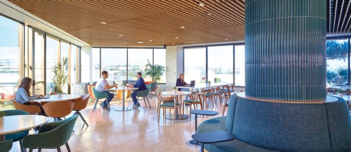 Spacious, sunlit lounge area with floor-to-ceiling windows featuring terrazzo flooring, mid-century modern furniture, and an acoustic fabric-wrapped column. Suspended wooden slats create a warm, textured ceiling, complementing the room's earth-toned color palette and offering a serene work and social environment.