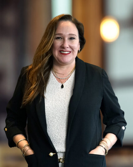 The image shows a woman with long dark hair and a beaming smile, wearing a black outfit with a patterned scarf and layered necklaces. The backdrop is softly focused with light tones, suggesting an open and contemporary workspace.