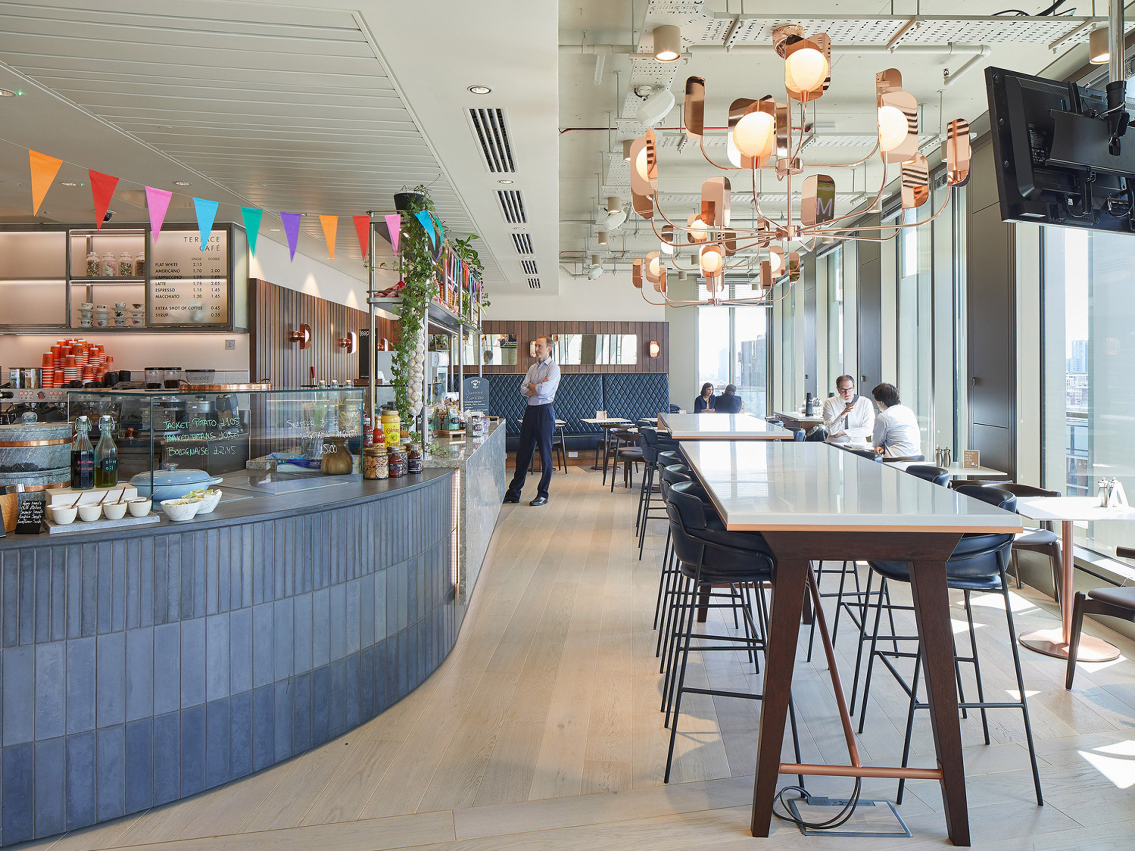 Open-plan cafe interior featuring a long counter with blue paneling, topped with an array of pastries and fruits. Pendant lights with a warm glow dangle above, and a communal high table occupies the foreground, surrounded by stools. Patrons converse casually by floor-to-ceiling windows offering ample natural light.