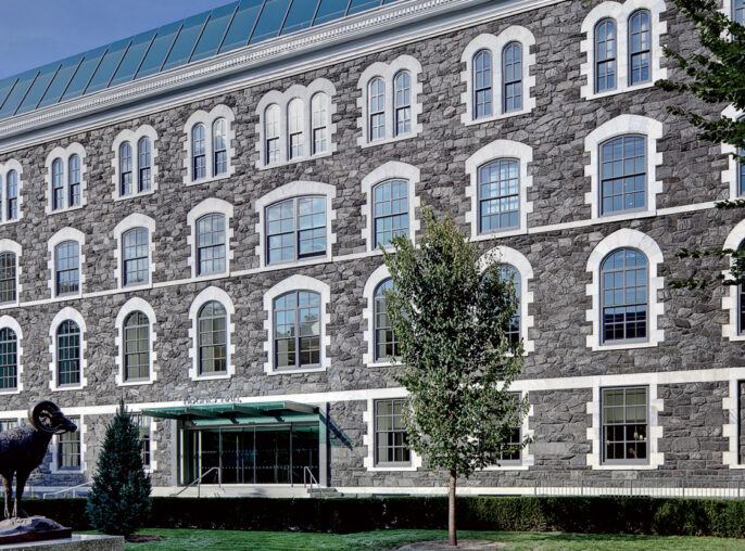 Exterior of a renovated historical building, showcasing a well-preserved stone facade, symmetrically aligned arched windows, and a modern glass rooftop addition that contrasts with the traditional architecture below. A sculptural art piece adorns the manicured front lawn.