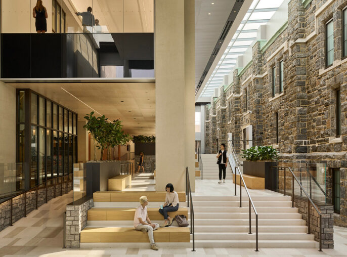 Modern interior juxtaposing historical stone walls with glass partitions, sleek black metal frames, and a sunlit atrium. Elegantly crafted stairs and tiered seating areas facilitate flow and interaction, complemented by geometric floor patterns that enhance the space's contemporary yet timeless ambiance.