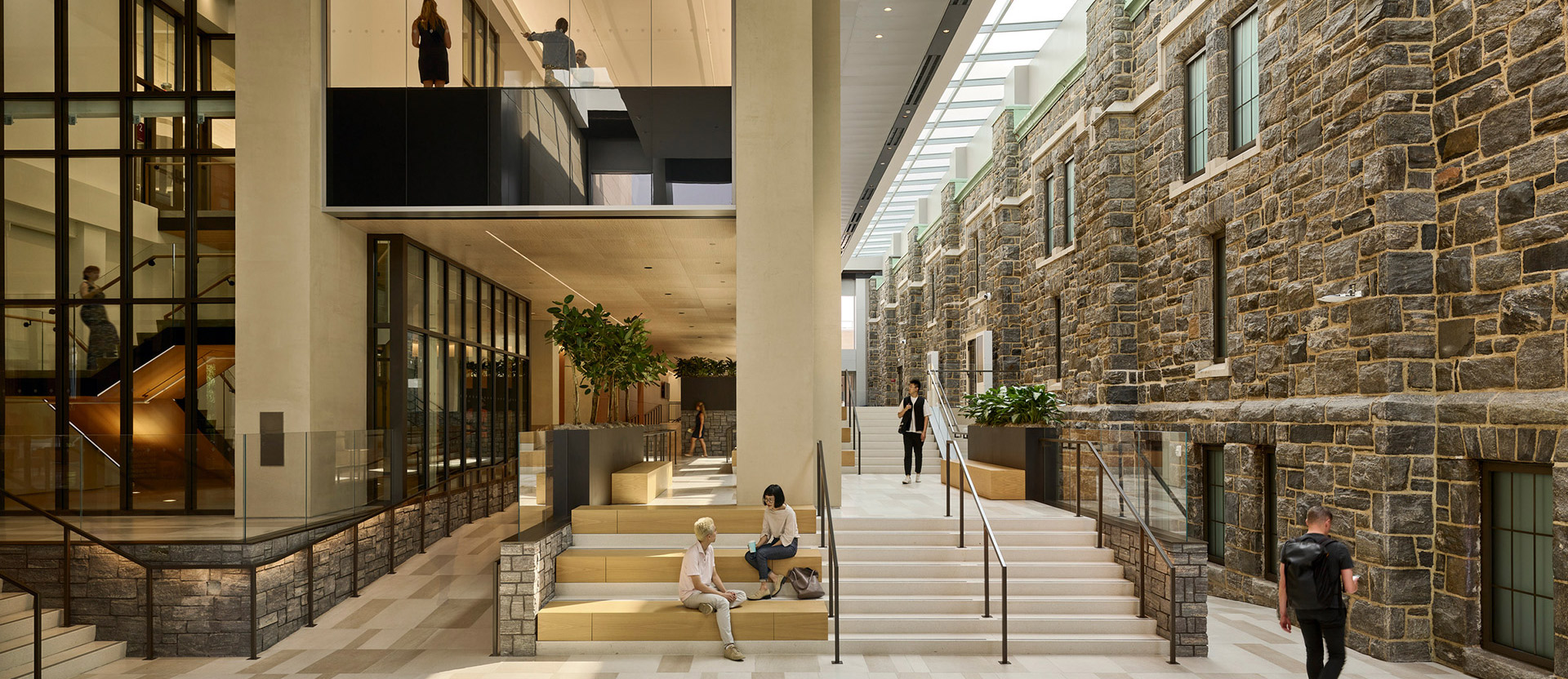 Modern interior juxtaposing historical stone walls with glass partitions, sleek black metal frames, and a sunlit atrium. Elegantly crafted stairs and tiered seating areas facilitate flow and interaction, complemented by geometric floor patterns that enhance the space's contemporary yet timeless ambiance.