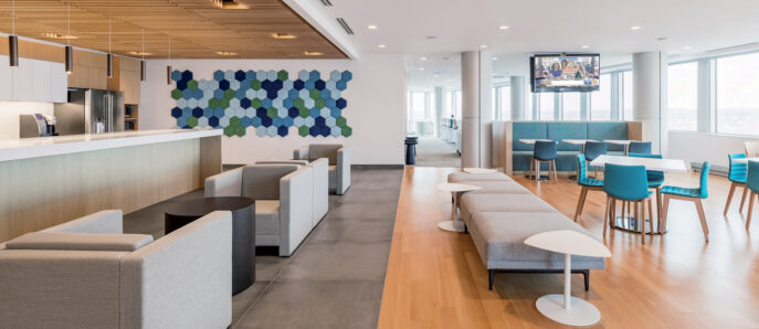 Modern office breakroom featuring natural light, sleek white surfaces, wooden slats ceiling detail, and pops of color from blue upholstered furniture and hexagonal wall art.