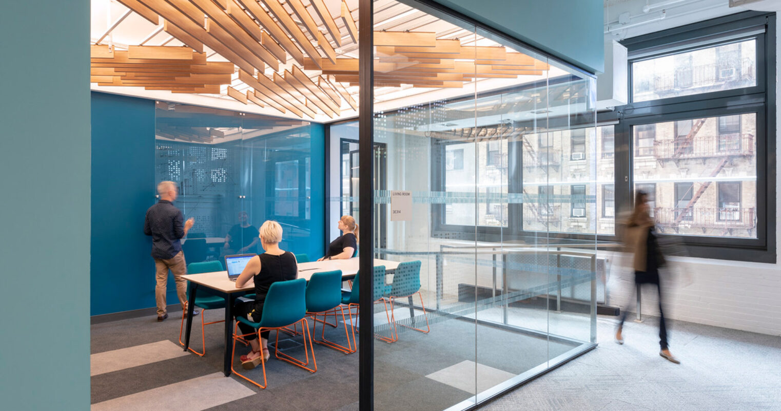 Contemporary office space featuring glass walls, a bold blue accent wall, and suspended wooden slats. Occupants seated at a table with modern chairs on a patterned gray carpet, engaging in a meeting, with natural light streaming through large windows.