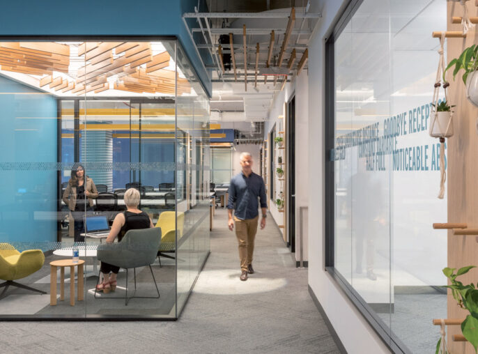 Modern open-plan office space featuring natural wood accents, hanging greenery, and glass partitions fostering transparency and collaboration, complemented by pops of blue and yellow in the seating areas.