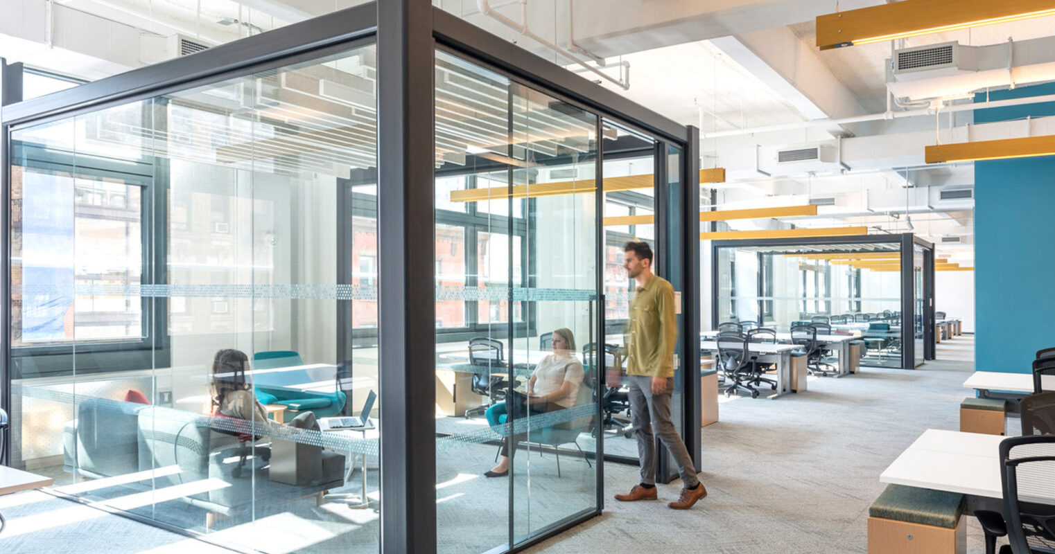 Bright, modern office space featuring glass partition walls creating a semi-private meeting area. Sleek, black frames contrast with the warm wood tones and pops of blue and yellow in the open-plan workspace beyond. Natural light floods the space, enhancing the airy ambiance.