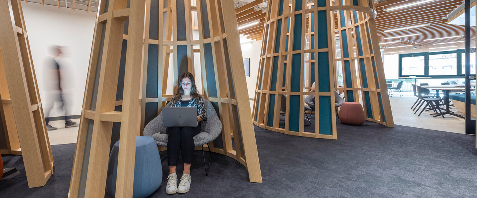 Modern office interior featuring distinctive wooden, tree-like structures creating semi-private seating areas. A person works on a laptop, seated on a blue pouf, amidst these organic design elements, which harmonize with the industrial-chic open-plan workspace.