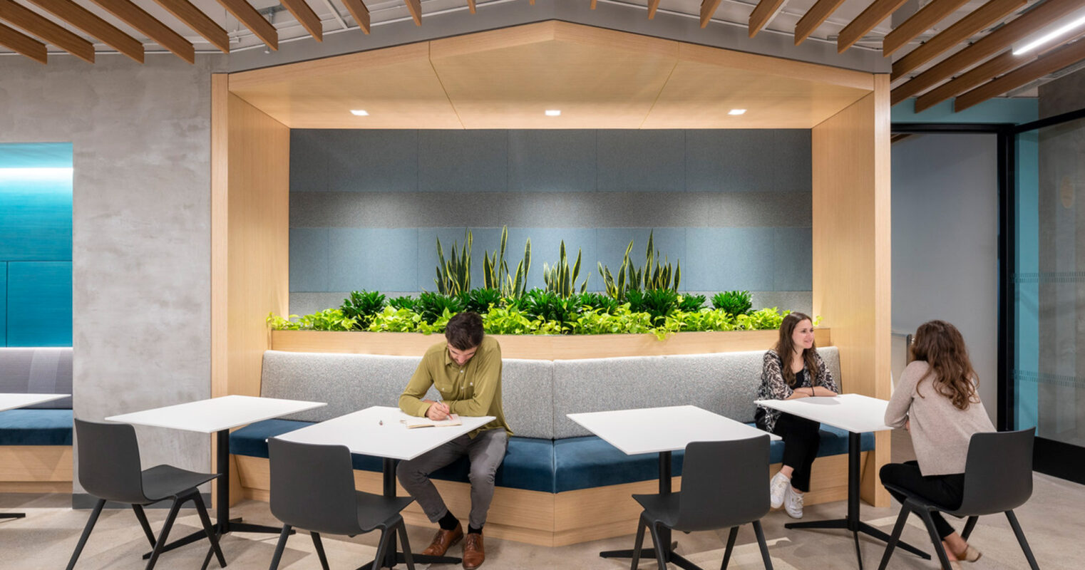 Modern office break area featuring a central plant bed with lush greenery, surrounded by high-backed banquettes and minimalist tables, under a ceiling adorned with warm wood slats and integrated lighting, providing a tranquil space for relaxation and informal meetings.